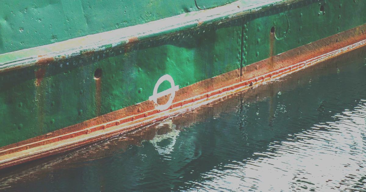 A green riveted hull at its waterline, with the white load-line disc and the rust boot-topping stripe above dark rippling water.